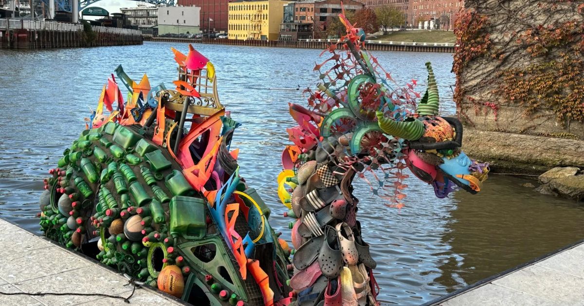 Large sculptures made from plastic waste sit beside a river, drawing attention to plastic pollution along an urban waterfront.