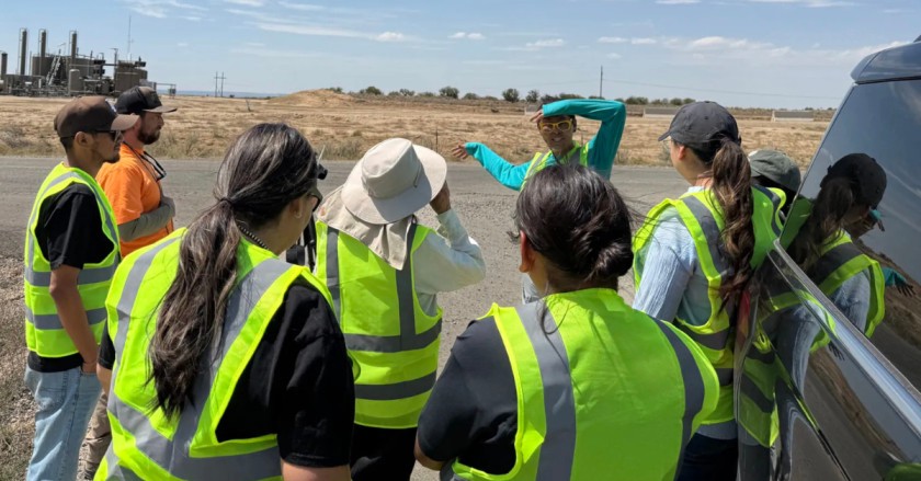 Group wearing neon safety vests gathers roadside as a guide gestures toward an industrial facility in the distance.