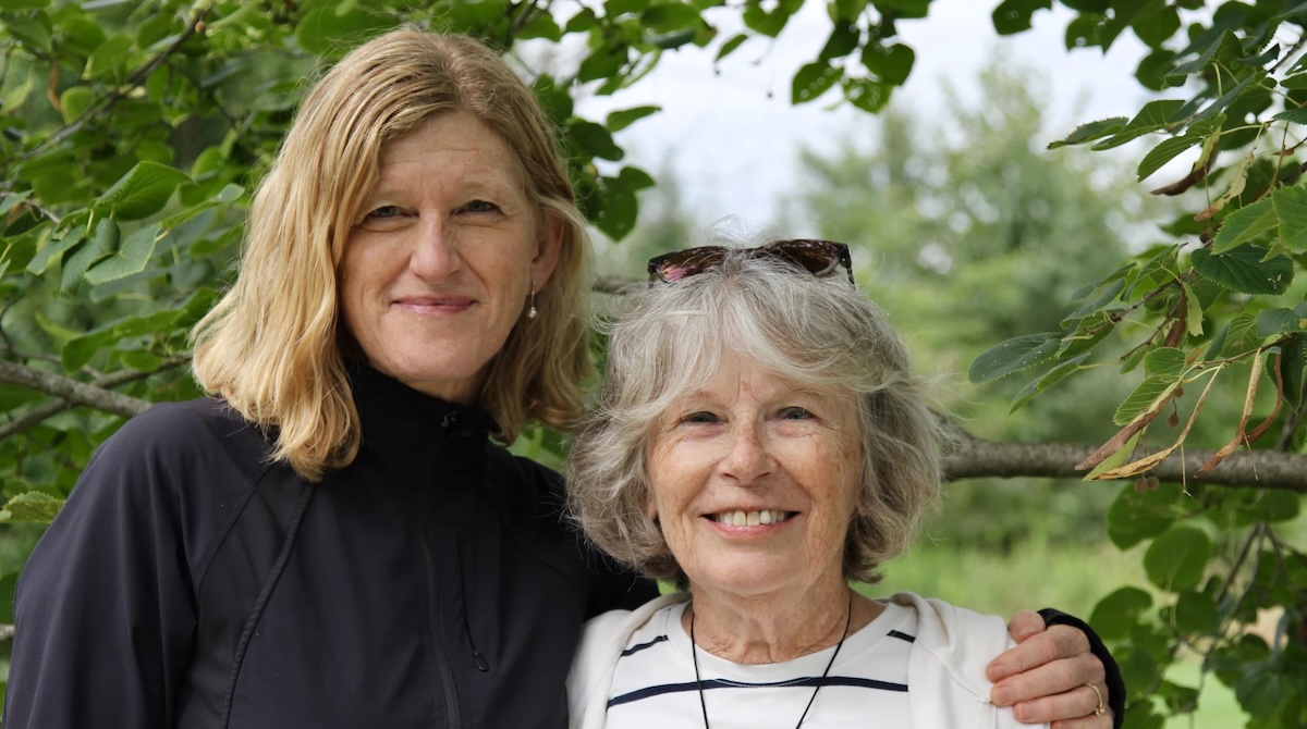 Kathy, a white woman with short, white hair and crinkly laugh lines, smiles at the camera. Ginny, who is half a head taller and has shoulder-length blond hair, has her arm around Kathy's shoulder. The braches of a beech tree spread to fill the frame behind the two women.