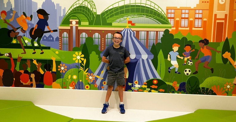 A boy in a blue shirt smiling and standing in front of a mural at the hospital