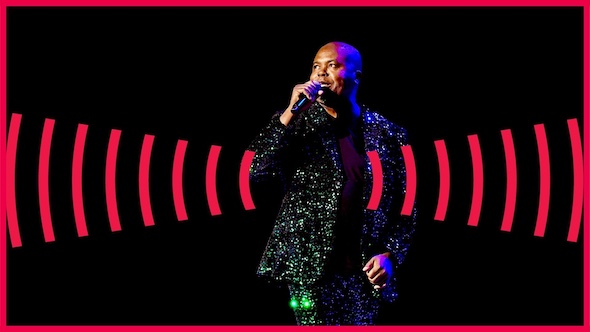 Spooked host Glynn Washington speaks into a mic on stage while wearing a sparkly green and purple suit.