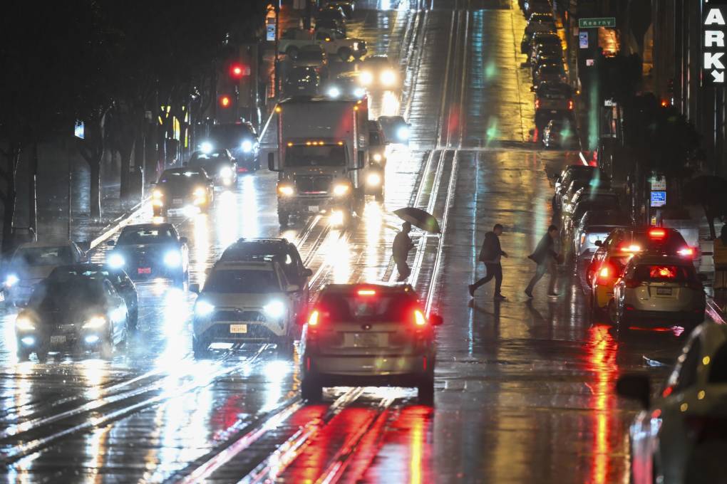 A small group of people, one using an umbrella, cross a busy street after dark as the pavement glitters with light reflecting off the wet road