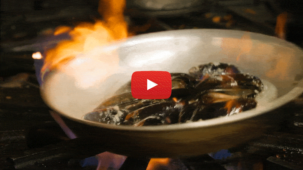 Moving image of mussels cooking on a pan with flames surrounding the sides.