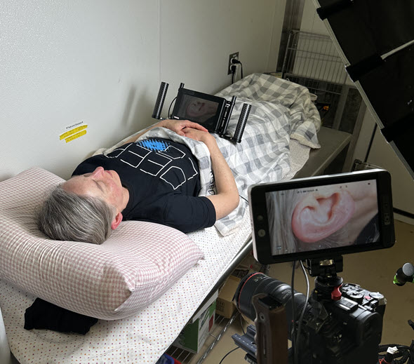 A filming monitor rests on Gabriela's stomach so she can see what’s being filmed. A close-up image of her ear is on a second monitor.