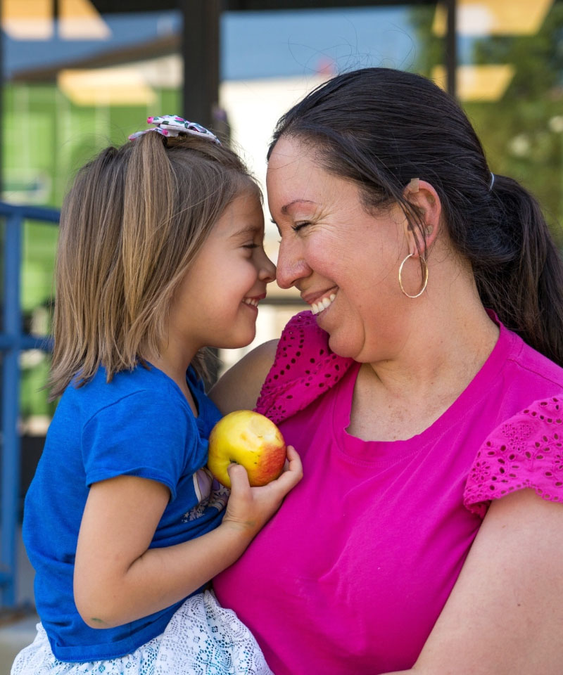 A joyful woman in a pink blouse and young girl in a blue shirt share a tender moment, smiling with noses touching, as the girl holds a yellow apple.
