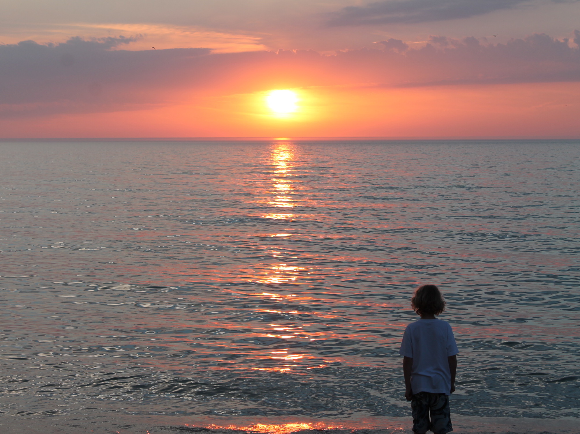 A young boy stands on the shoreline at sunrise.